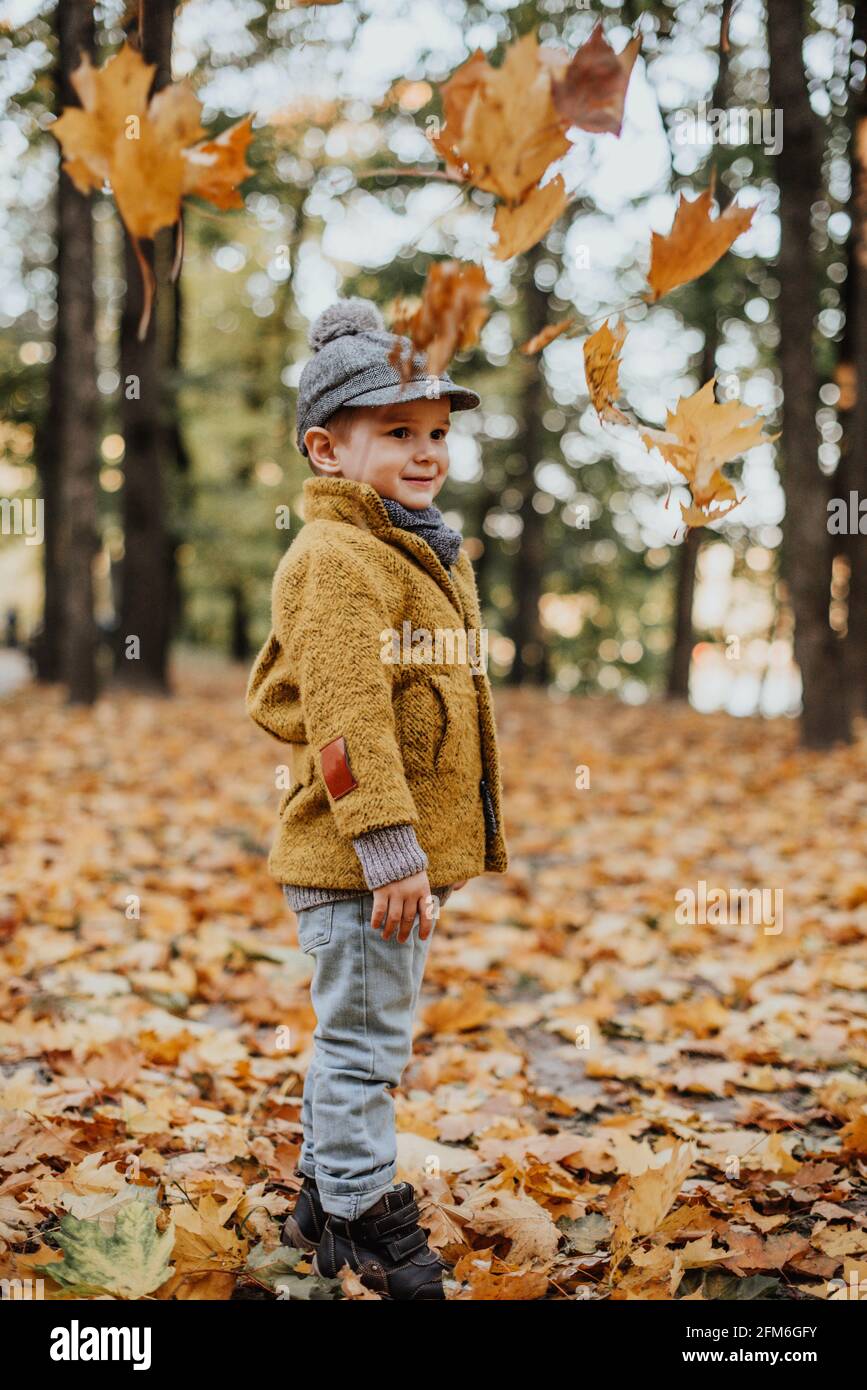 Garçon élégant s'amusant dans le parc de la ville d'automne. Joyeux gamin  jouant des feuilles mortes. Mode pour enfants. Garçon portant un manteau,  une casquette et une écharpe jaune tendance. Jeune garçon