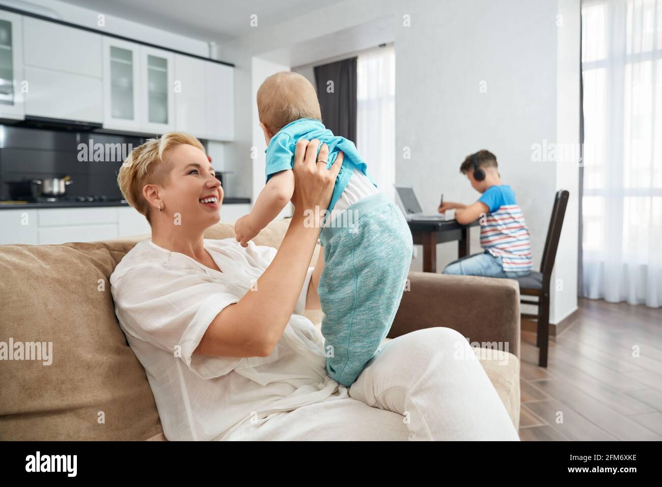 Femme souriante aux cheveux blonds courts prenant soin de son petit fils tandis que le fils aîné s'asseyant à la table de cuisine et étudiant sur un ordinateur portable. Concept de famille et de technologie. Banque D'Images