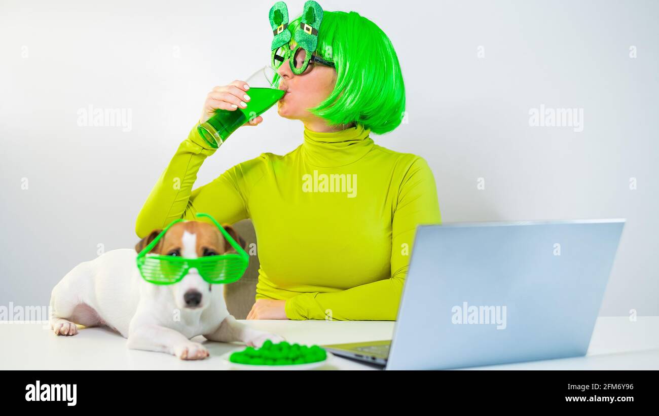 Une jeune femme dans une perruque verte et des verres gaies boit de la bière et des piqûres de noix glacées. Une fille s'assoit avec un chien à une table et célèbre le jour de St patrick Banque D'Images