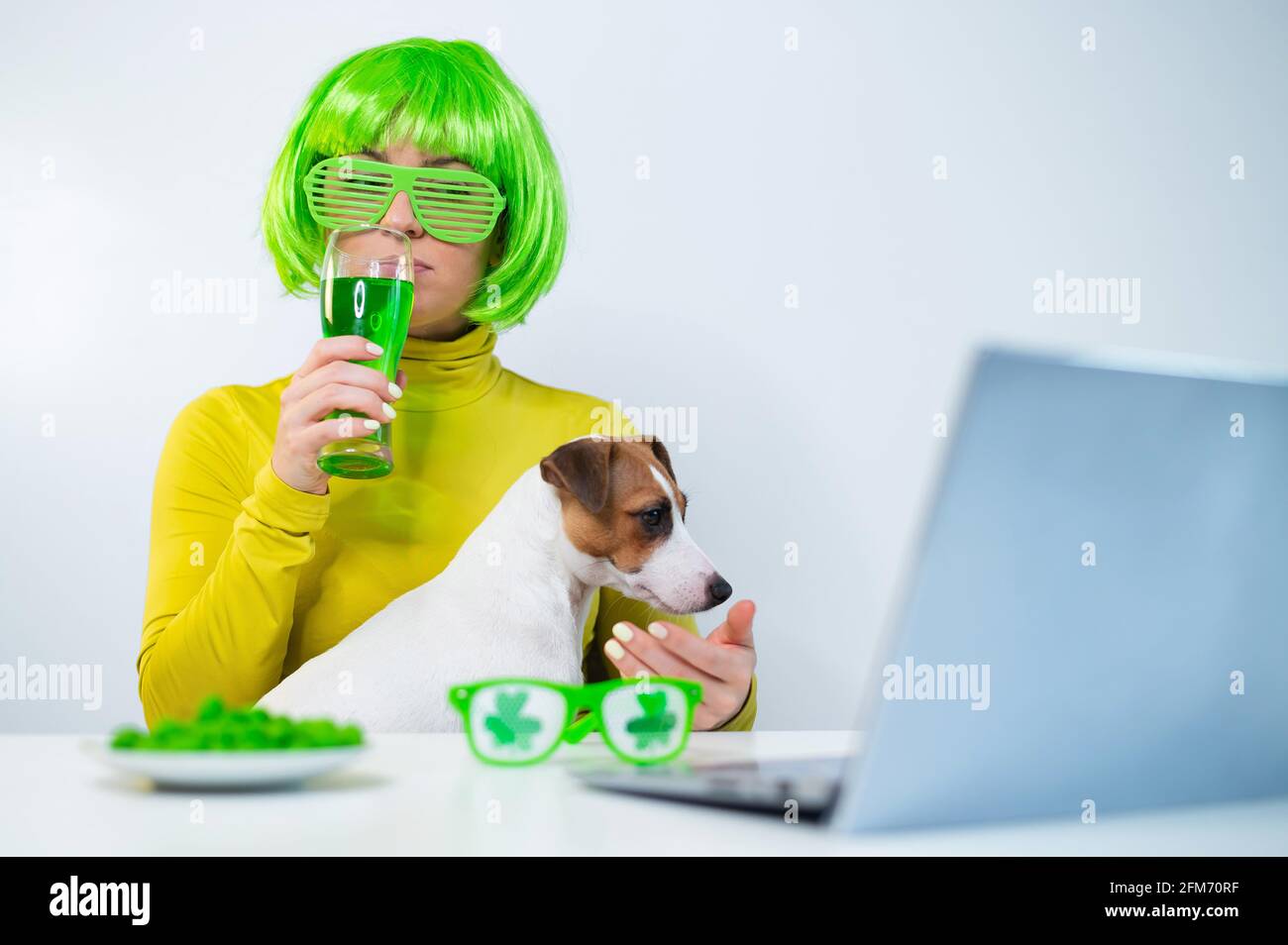 Une jeune femme dans une perruque verte et des verres gaies boit de la bière et des piqûres de noix glacées. Une fille s'assoit avec un chien à une table et célèbre le jour de St patrick Banque D'Images