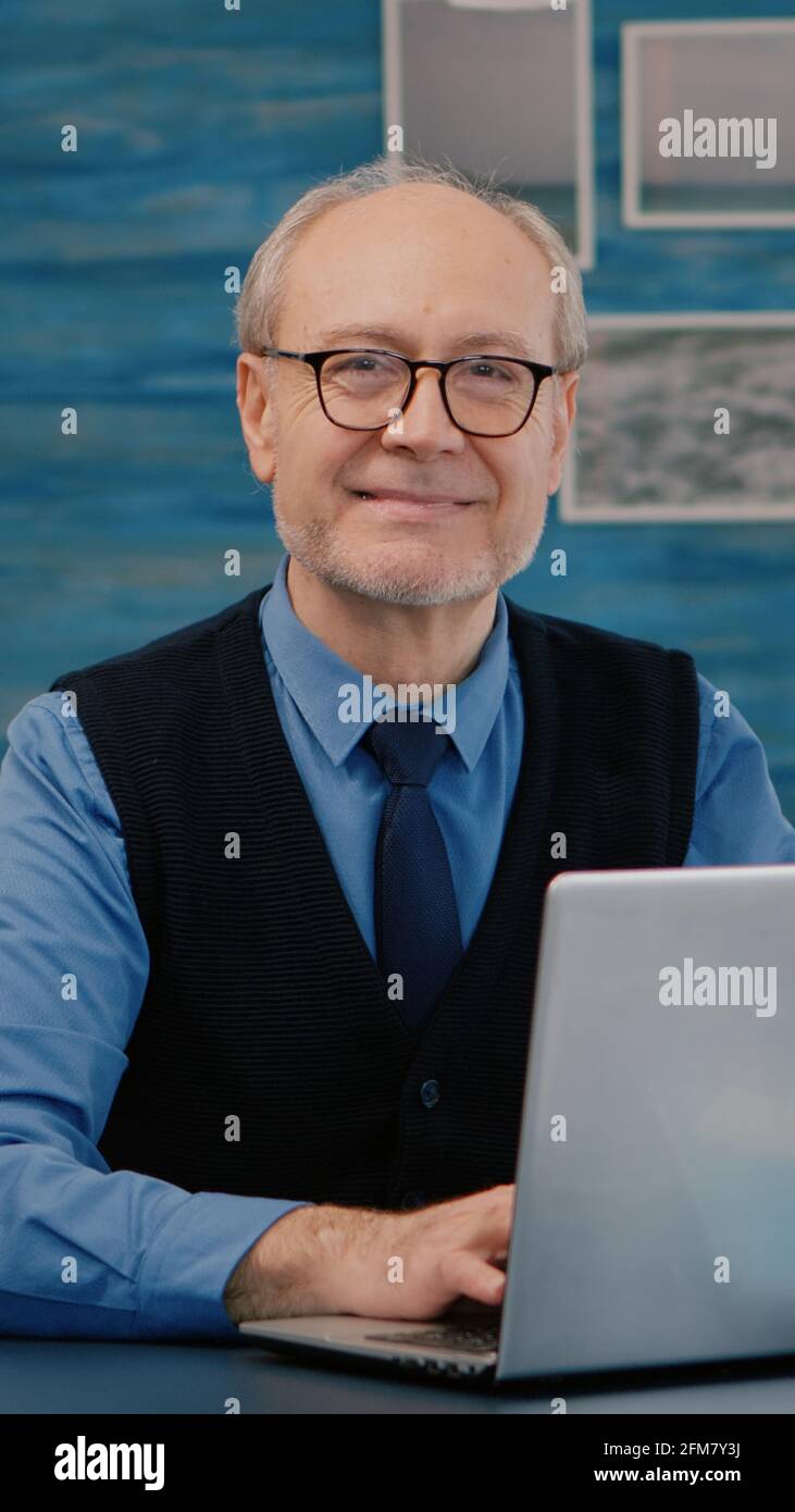 Un directeur à la retraite assis à la réception devant l'appareil photo souriant après avoir tapé sur un ordinateur portable à la maison pendant que la femme principale lisait un livre en arrière-plan. Homme préparé pour la réunion en ligne à l'aide de gadgets moden Banque D'Images