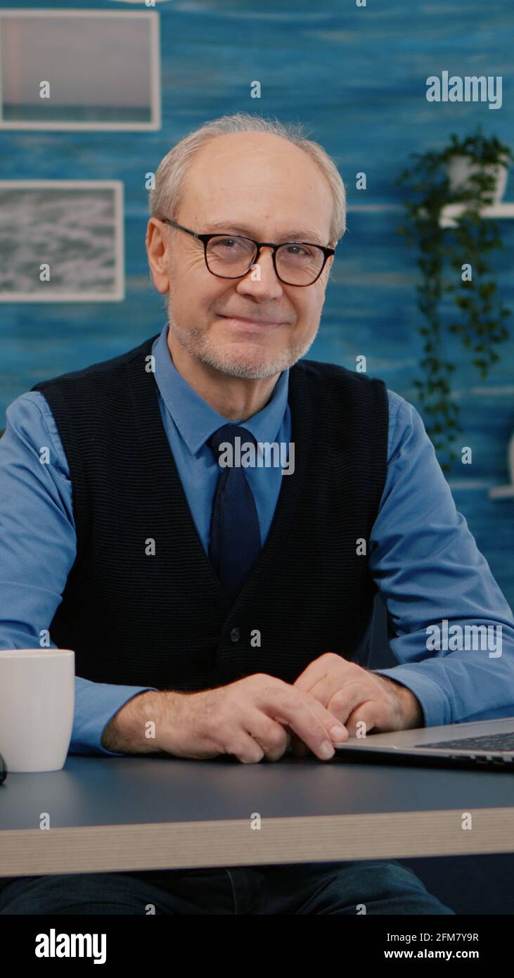 Homme senior regardant la caméra souriant assis sur le lieu de travail au bureau, travaillant de la maison tandis que la vieille femme regardait la télévision en arrière-plan. Personne d'affaires à la retraite préparée pour une réunion en ligne à l'aide de gadgets moden Banque D'Images