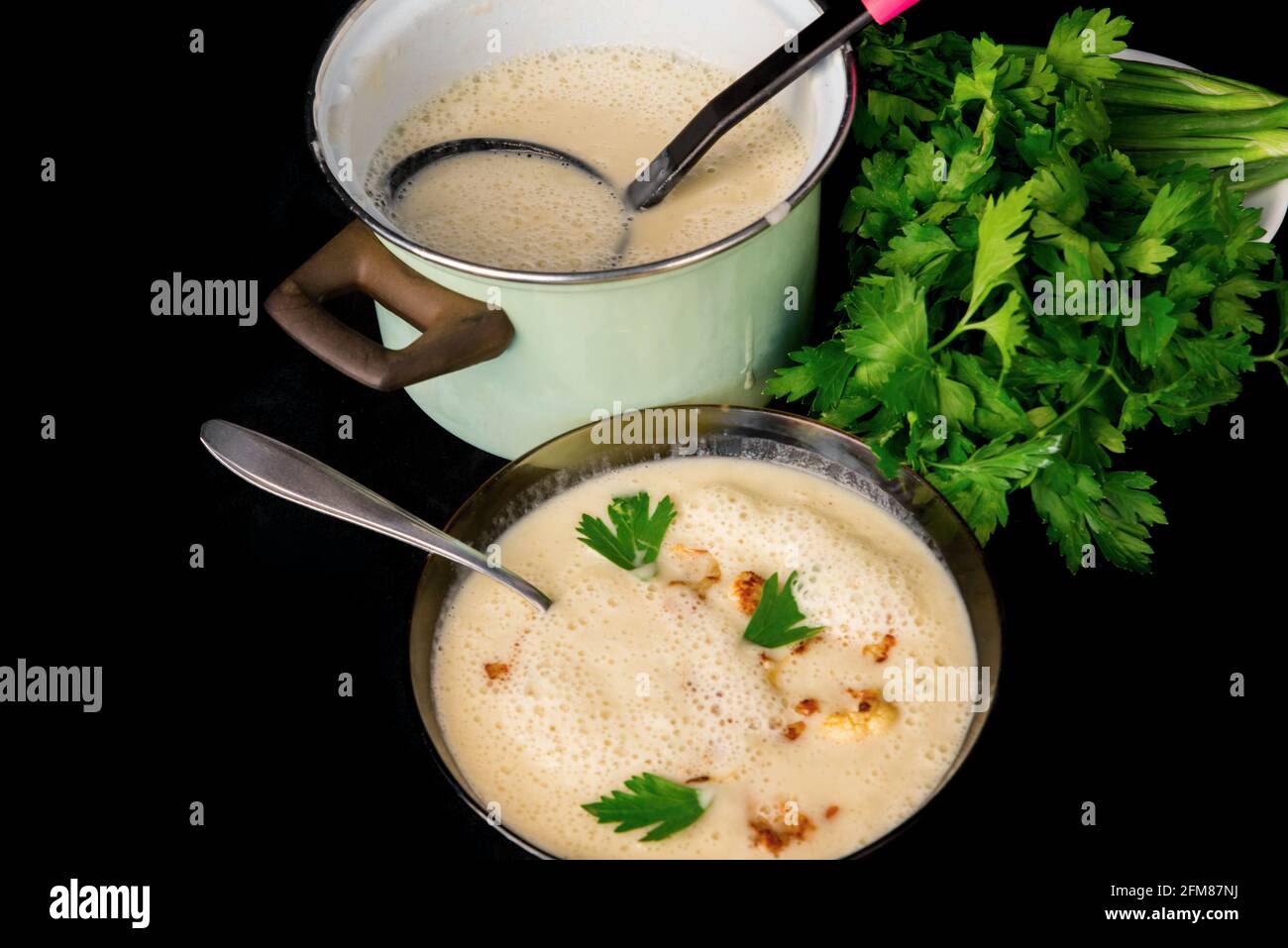 Soupe crémeuse de chou-fleur dans une assiette et dans une casserole verte avec louche et bouquet de persil vert sur fond noir. Banque D'Images Soupe crémeuse de chou-fleur dans une assiette et dans une casserole verte avec louche et bouquet de persil vert sur fond noir. Banque D'Images