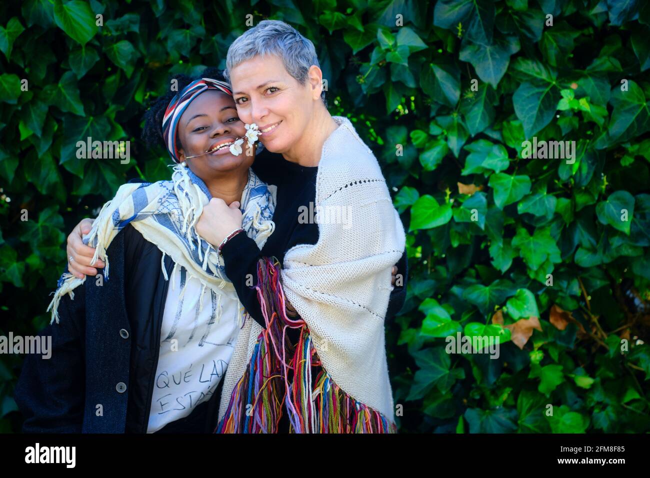 Femme caucasienne mature et jeune femme africaine embrassant dans un jardin avec un fond de feuilles. Banque D'Images