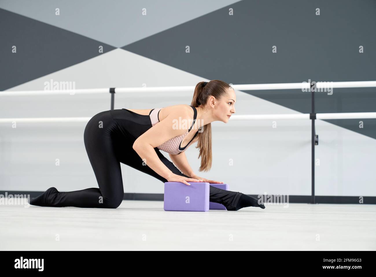 Vue latérale d'une femme à la coupe attrayante avec queue de cheval de châtaignier qui s'étend sur la jambe avec de petits blocs violets sur le sol. Jolie danseuse pratiquant dans une salle vide. Étirement, gymnastique concept. Banque D'Images
