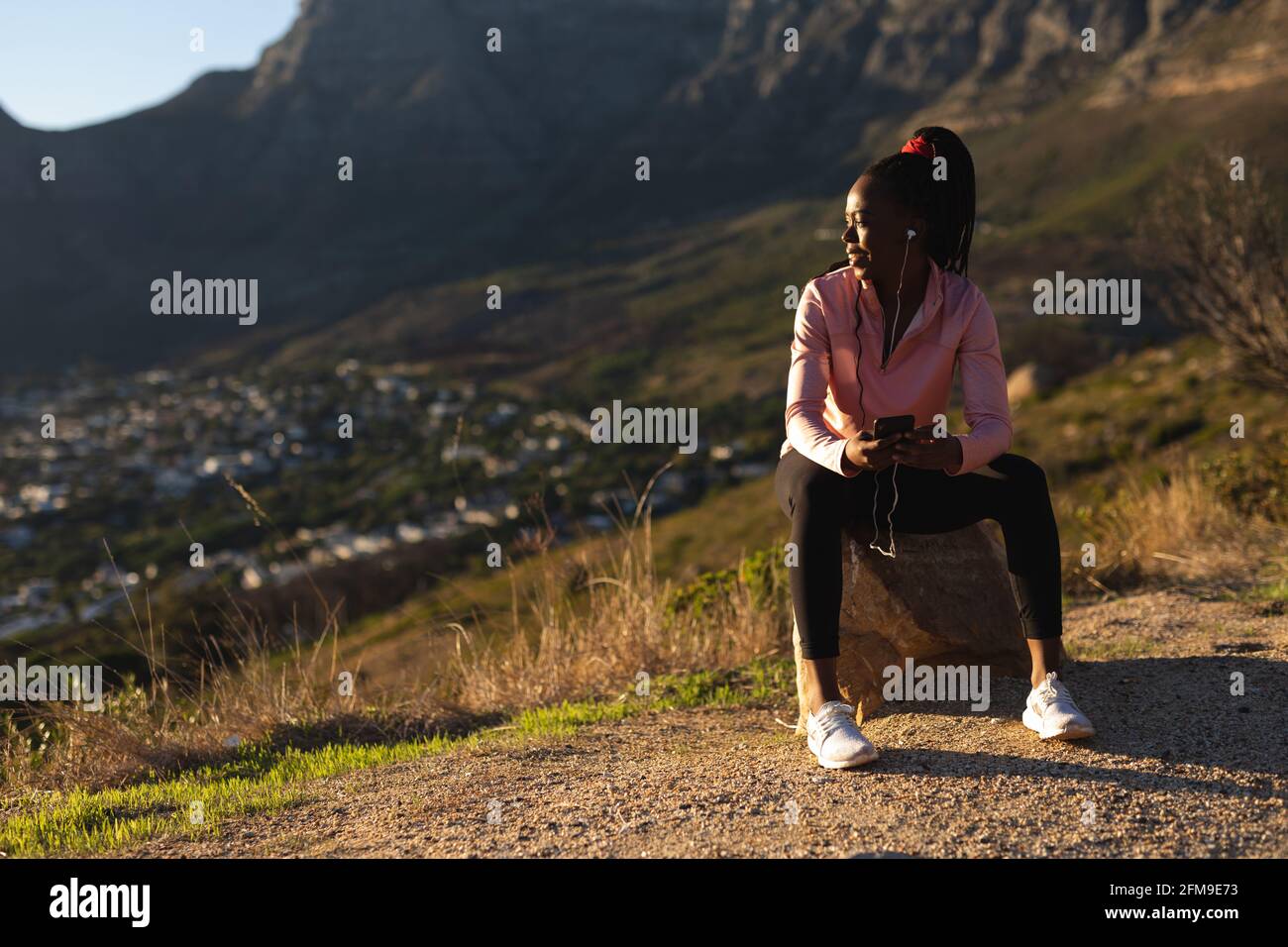 Femme afro-américaine prenant une pause dans l'exercice à l'extérieur, portant des écouteurs et tenant un smartphone Banque D'Images