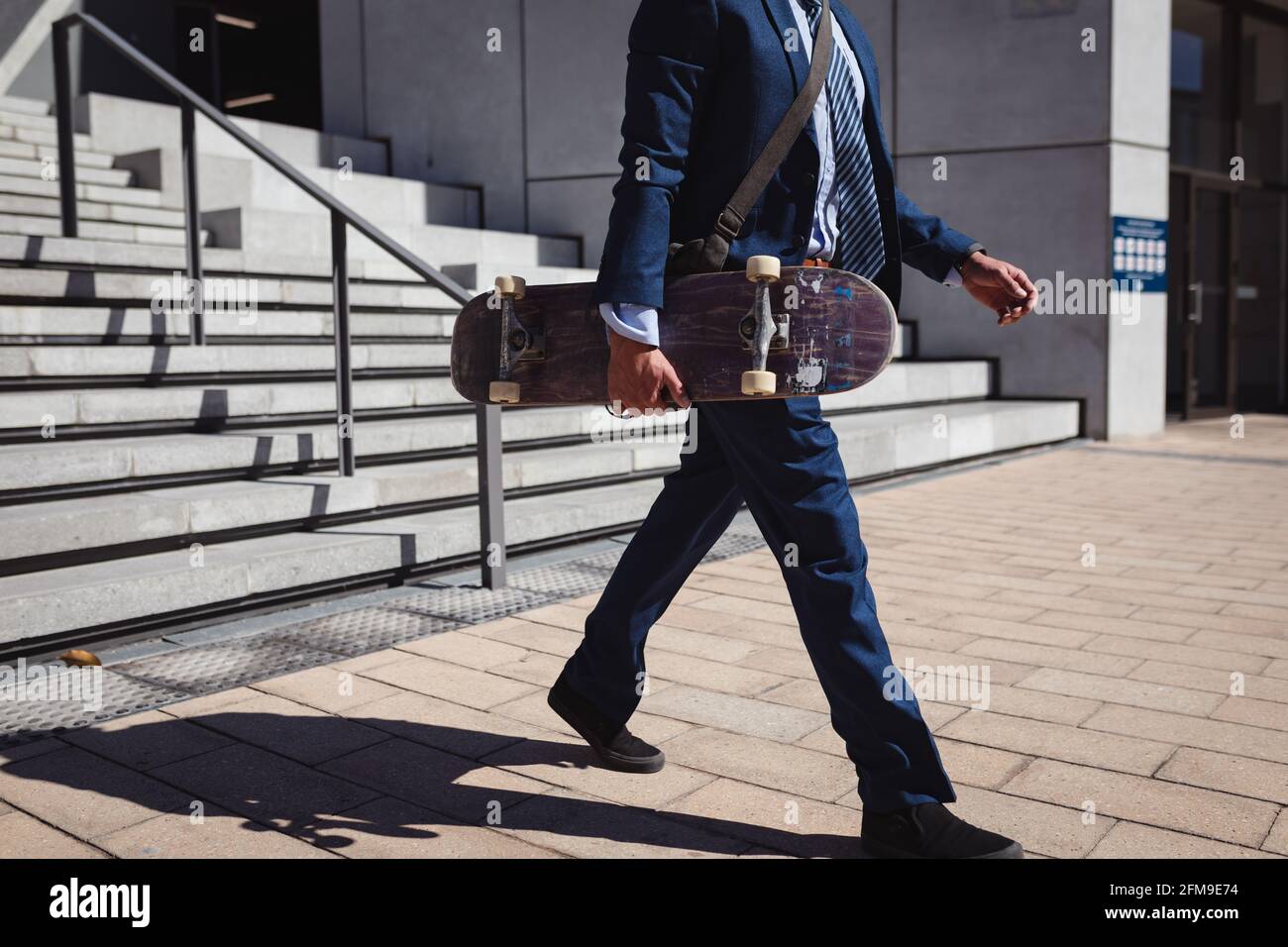 Bas de section d'homme d'affaires caucasien tenant le skateboard, marchant au soleil Banque D'Images