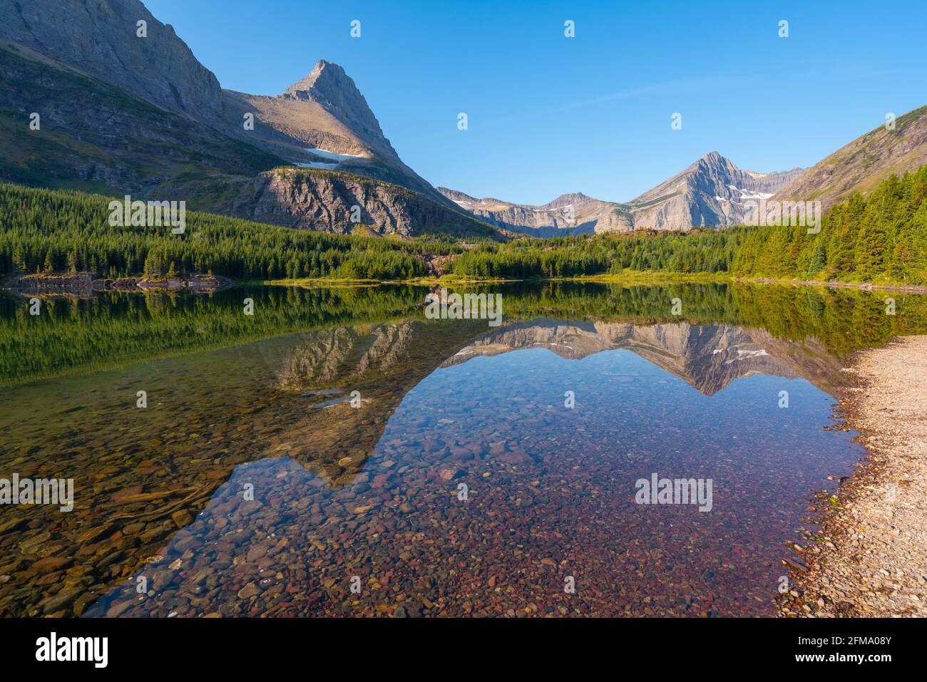 Journée ensoleillée au bord du lac Bullhead dans le parc national des Glaciers. Reflet de la chaîne de montagnes à la surface de l'eau. Galets rouges sur la plage. Randonnée à SwiftCurrent Banque D'Images