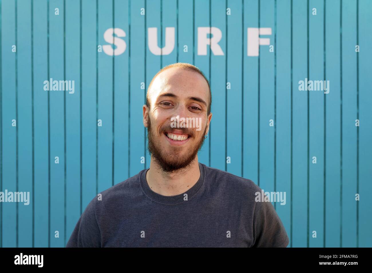 Le jeune homme heureux barbu sourit à la plage d'Isla Canela debout devant un mur en bois bleu avec le mot surf avec un rayon de soleil sur sa tête. Regardez Banque D'Images