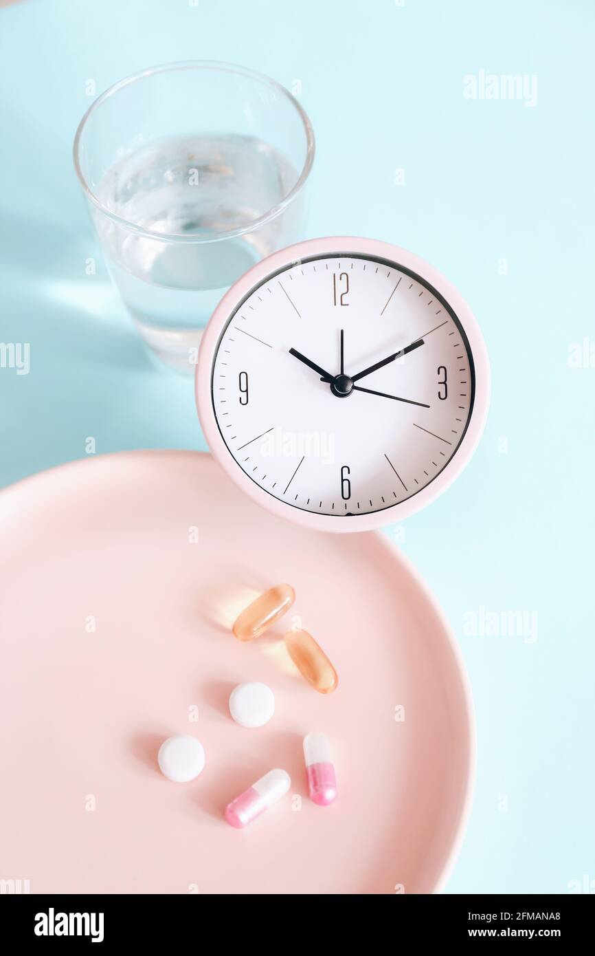 Il est temps de prendre des pilules. Horloge rose avec pilules et verre d'eau sur fond bleu. Vue de dessus. Pose à plat Banque D'Images