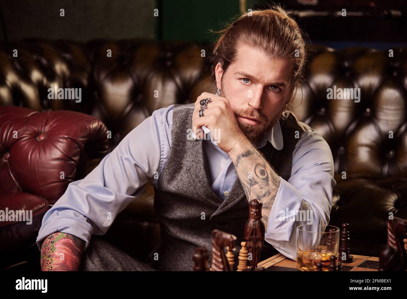 Jeune homme aux cheveux longs et tatouages au salon de coiffure avec jeu d'échecs Banque D'Images