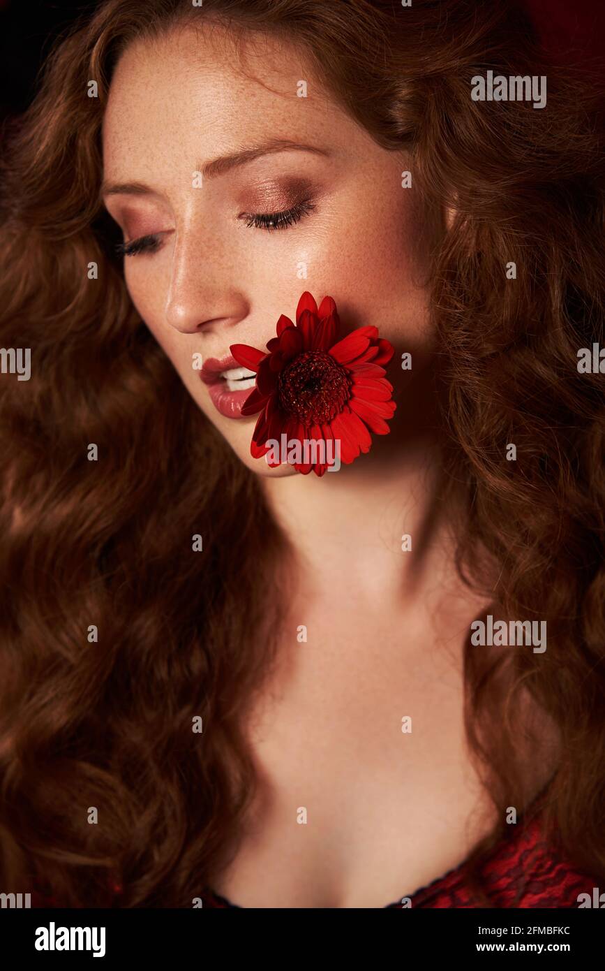 Jeune femme aux cheveux rouges luxuriants et aux taches de rousseur à la fleur dans le coin de sa bouche Banque D'Images