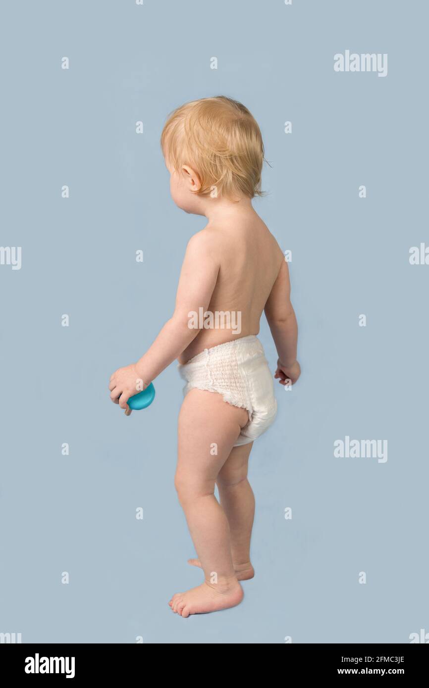 Une petite fille blonde de 1 an dans une couche blanche se tient sur un fond bleu. Vue de l'arrière Banque D'Images