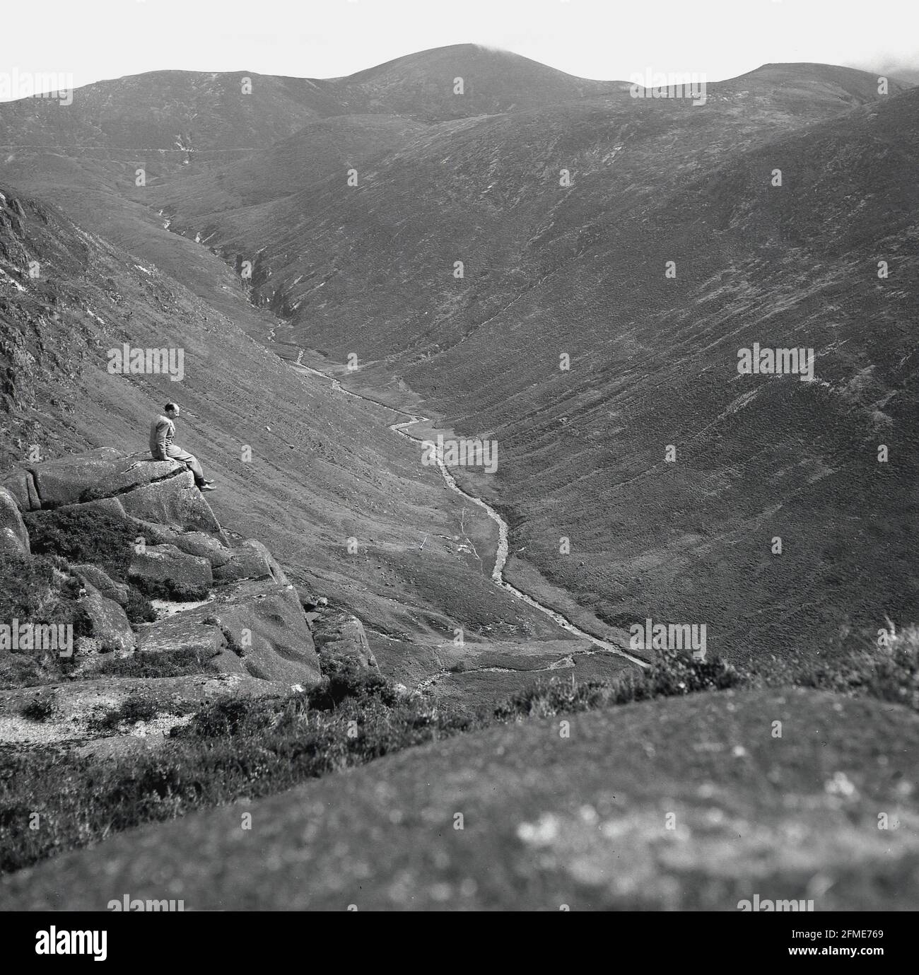 Années 1950, historique, homme en costume assis sur le rebord d'une roche en haut d'une colline dans les montagnes Antrim, surplombant une vallée profonde ou glen Below, Co Antrim, Irlande du Nord. Banque D'Images