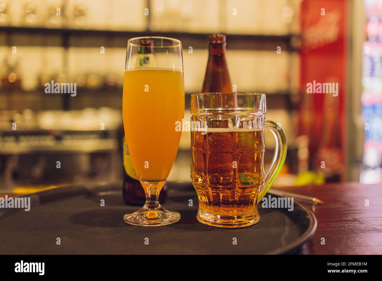 Gros plan sur une grande pinte remplie de bière de blé non filtrée, devant  3 verres de bière et de cidre, avec une femme en arrière-plan, sur une  table en bois à