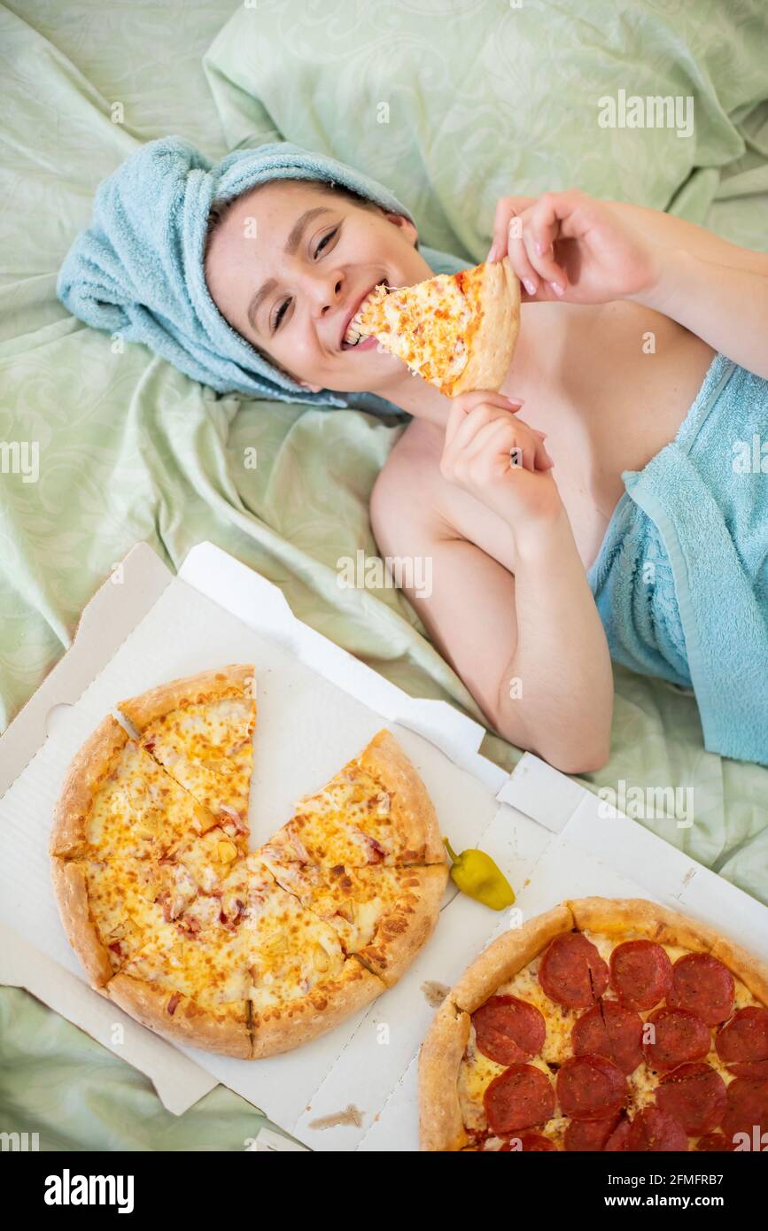 Une jolie fille avec une serviette sur sa tête mange de la pizza au lit.  Jeune femme mangeant de la pizza au lit. La vie est un plaisir, le corps  positif. J'adore