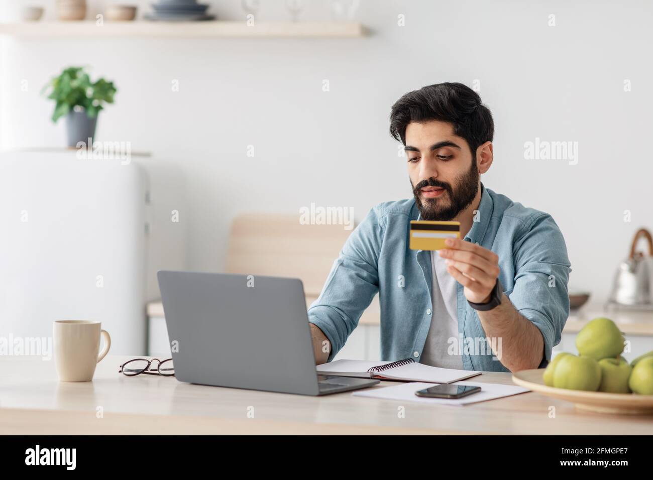 Homme arabe effectuant des paiements en ligne avec ordinateur portable et carte de crédit, assis dans kithcen à la maison, espace vide Banque D'Images