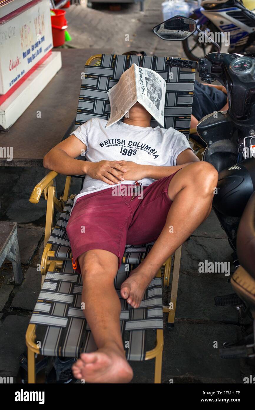 Un homme vietnamien allongé sur une chaise longue dort avec journal couvrant la face sur le trottoir, Hoi an, Vietnam Banque D'Images