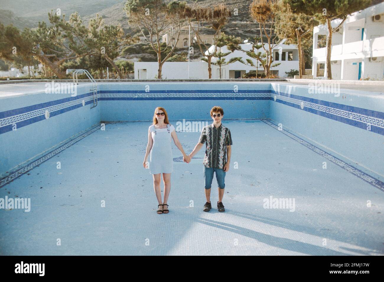 Ensemble corps jeune couple calme dans des tenues décontractées tenant les mains et regarder la caméra en se tenant dans la piscine sans eau dans une station ensoleillée Banque D'Images