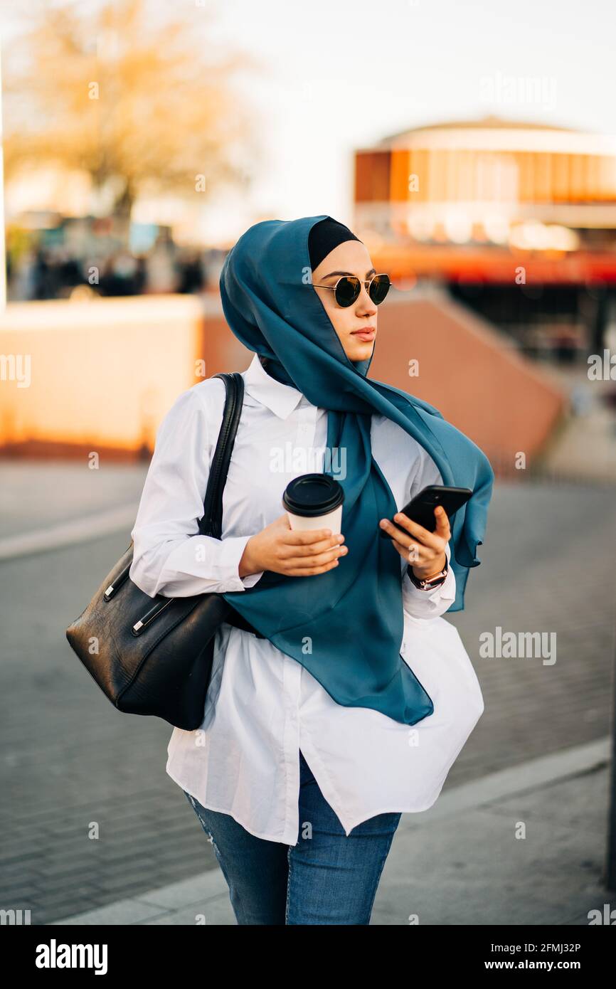 Femme ethnique en foulard et lunettes de soleil élégantes marchant avec des  plats à emporter prenez un verre dans la rue, tenez votre smartphone et  regardez loin Photo Stock - Alamy