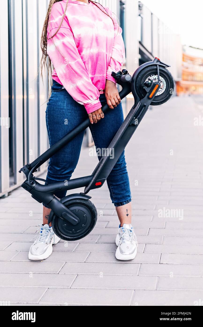 Crop anonyme femme en tenue décontractée debout avec scooter moderne sur une passerelle carrelée de la ville Banque D'Images