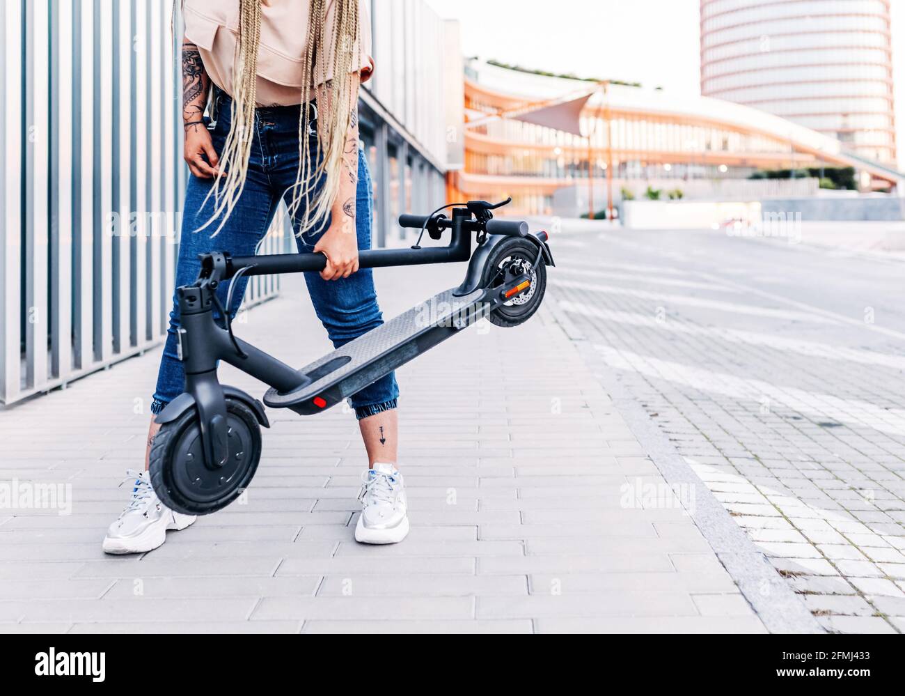 Crop anonyme femme en tenue décontractée debout avec scooter moderne sur une passerelle carrelée de la ville Banque D'Images