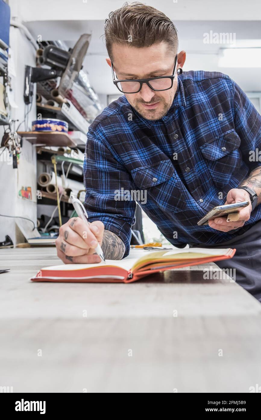 Homme barbu concentré artisan debout à l'établi et à l'écriture bloc-notes lors de l'utilisation du smartphone dans workshop Banque D'Images