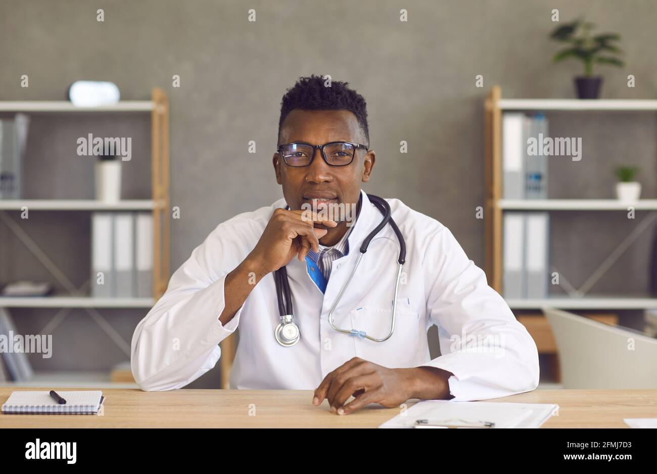 Portrait d'un jeune médecin noir en lunettes assis au bureau et écouter le patient Banque D'Images Portrait d'un jeune médecin noir en lunettes assis au bureau et écouter le patient Banque D'Images