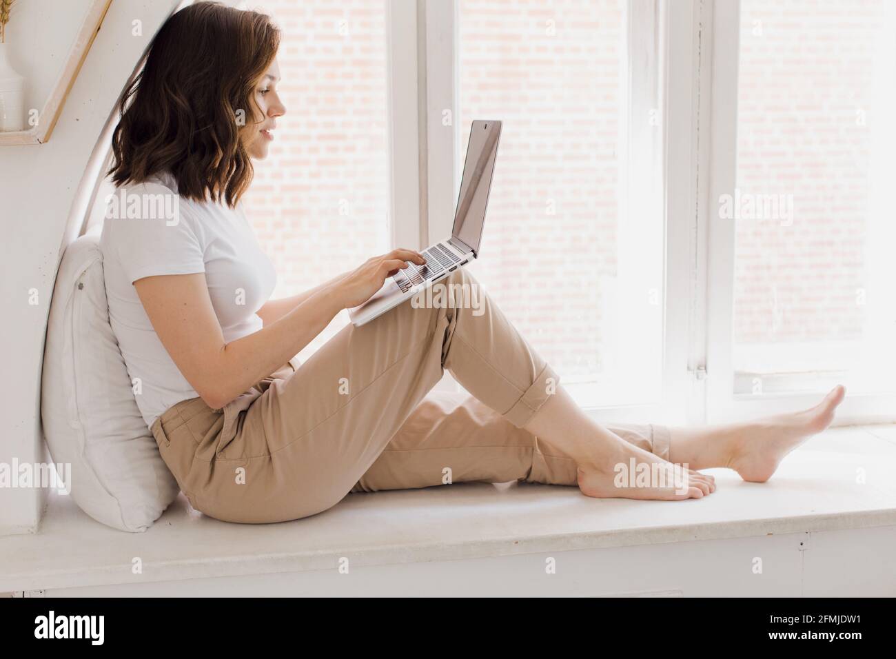 une jeune femme s'assoit sur le rebord de la fenêtre dans un confortable appartement lumineux et communique par vidéo. femme attrayante utilise un ordinateur portable pour son travail Banque D'Images