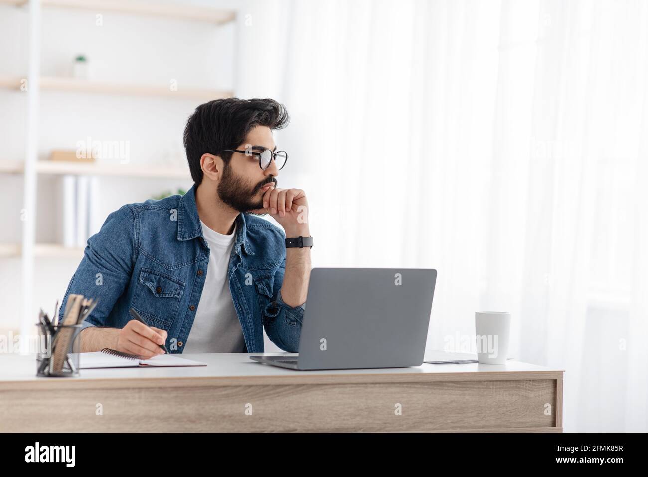 Un indépendant arabe réfléchi qui note des idées sur le bloc-notes, prend des notes sur le lieu de travail à domicile, planifie le programme de travail et le budget, s'assoit au bureau avec un ordinateur portable Banque D'Images