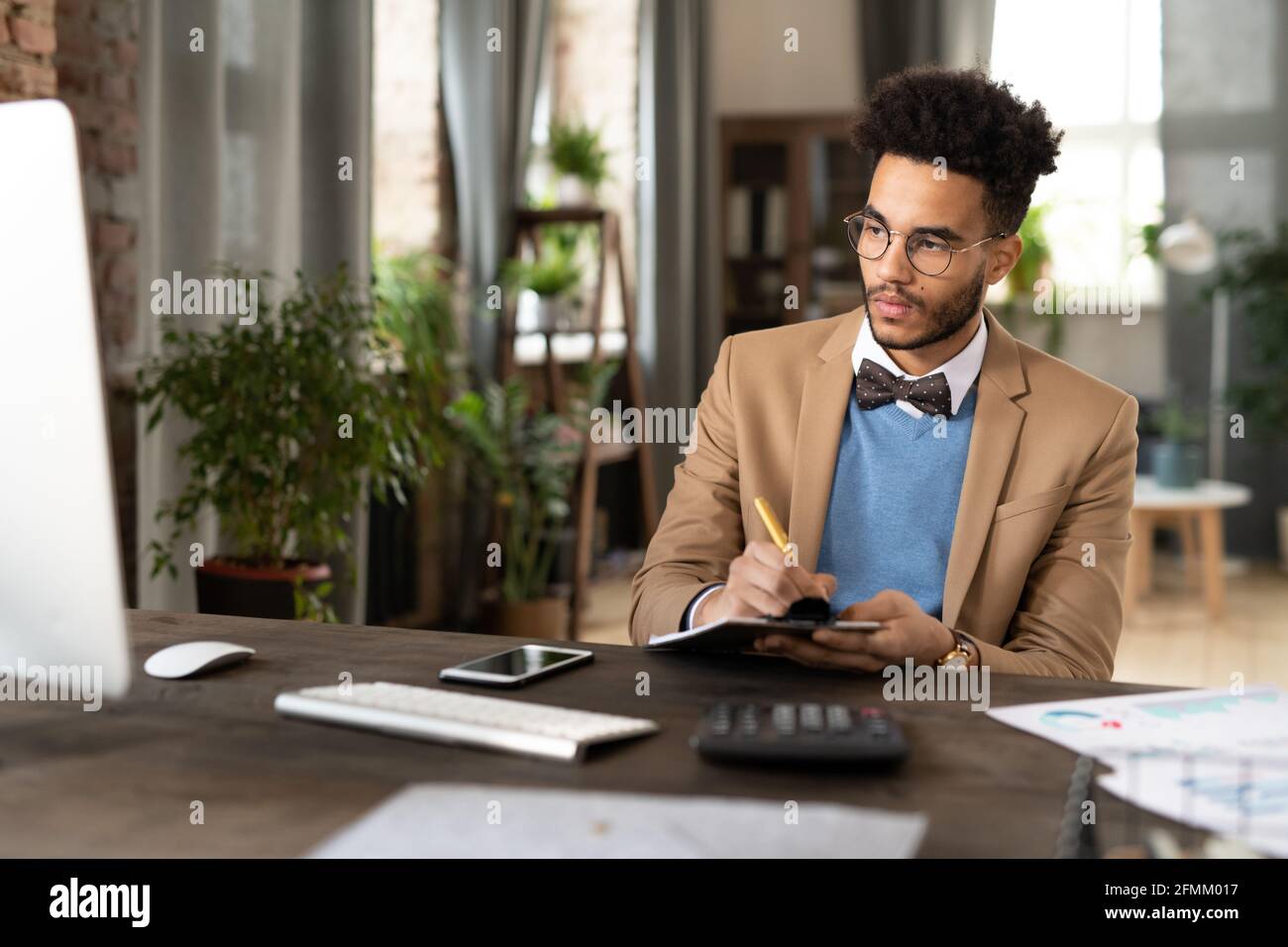 Jeune financier noir concentré avec barbe assis sur un bureau en bois avec la calculatrice et prendre des notes dans le bloc-notes lors de l'analyse des données sur l'ordinateur Banque D'Images
