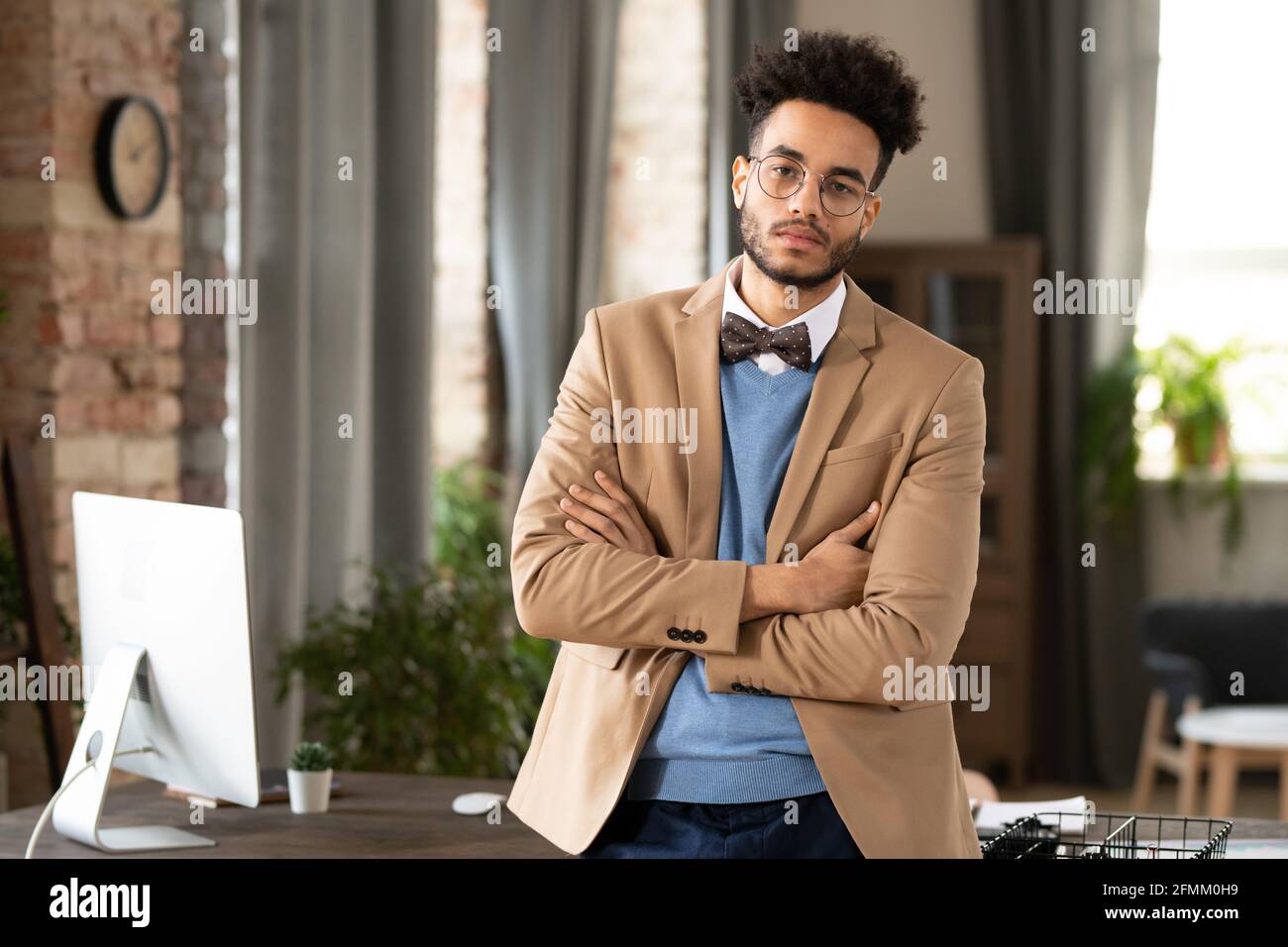 Portrait d'un Manager afro-américain confiant avec une barbe debout bras croisés contre la table avec un ordinateur moderne dans un bureau confortable Banque D'Images
