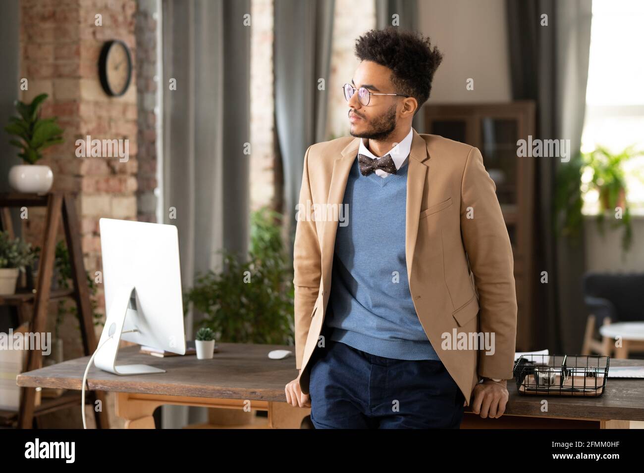 Jeune homme de course mixte sérieux en lunettes debout au bureau table avec ordinateur moderne et vue de côté Banque D'Images