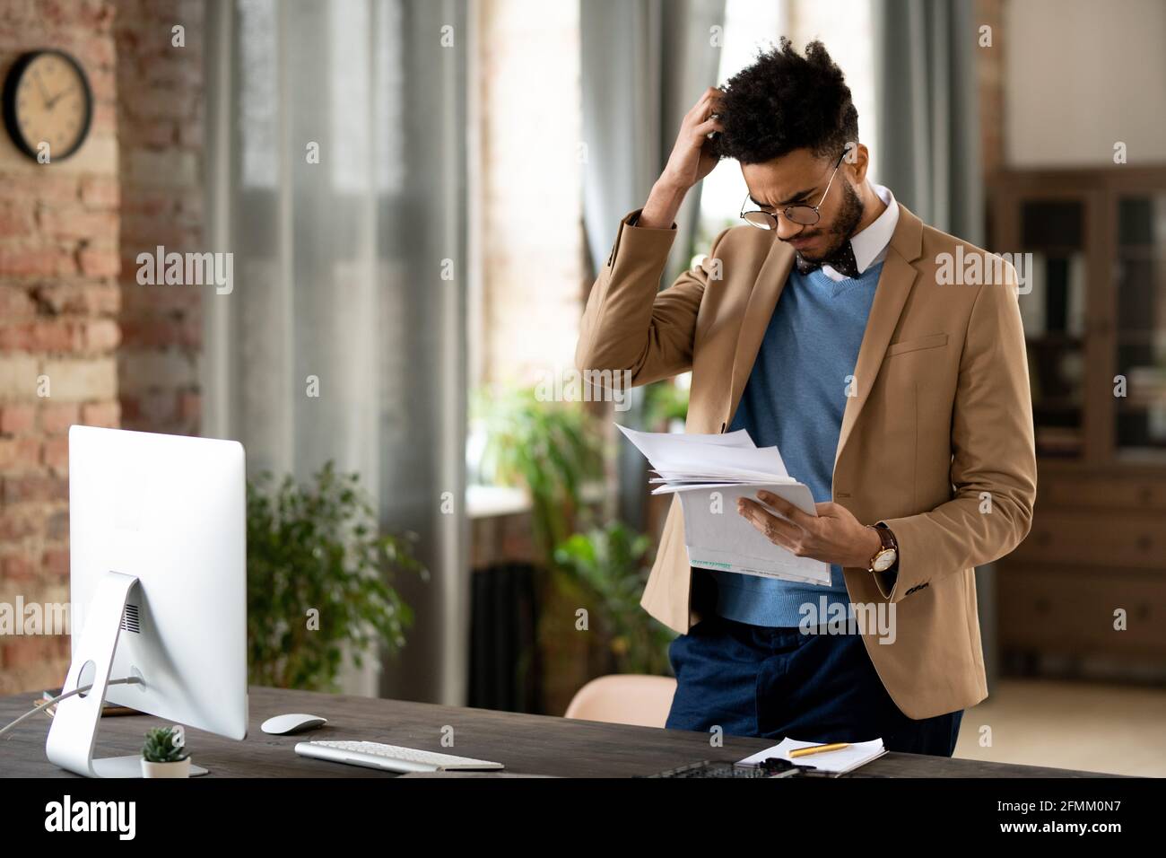 Jeune homme de course mixte confus en lunettes et veste debout au bureau avec ordinateur et en se grattant la tête tout en lisant des journaux Banque D'Images