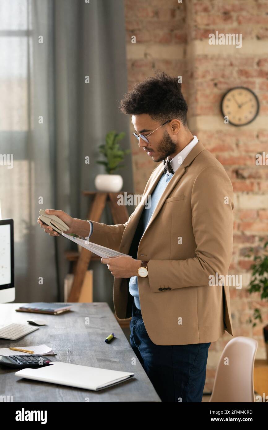 Jeune homme d'affaires sérieux et mixte avec une barbe debout à son bureau avec calculatrice et papiers d'agrafage au bureau Banque D'Images