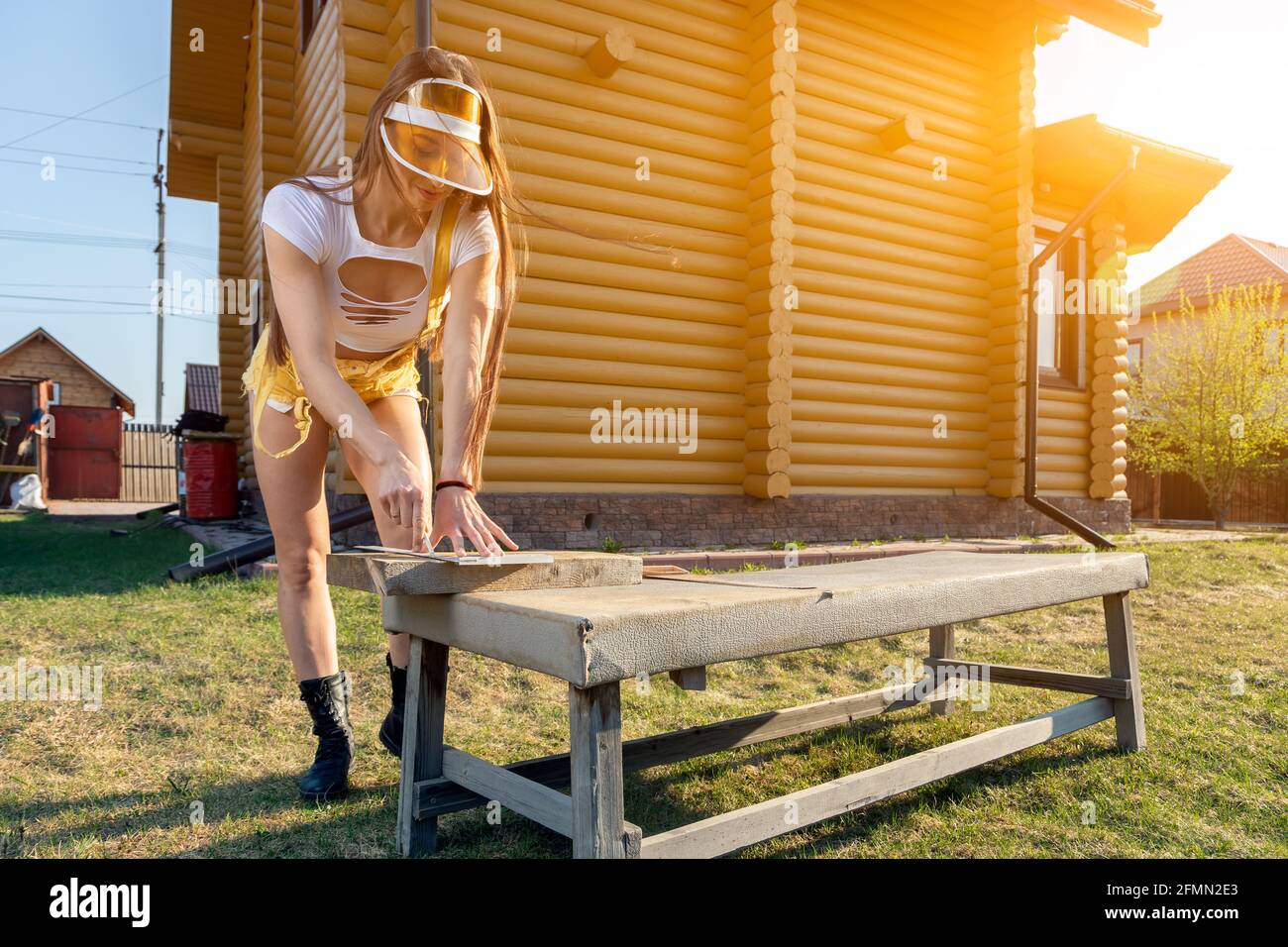 Jeune femme sportive en jeans et crop top est scier du bois dans une atmosphère rustique pendant une chaude journée d'été, dans le fond une maison en bois et une pelouse Banque D'Images