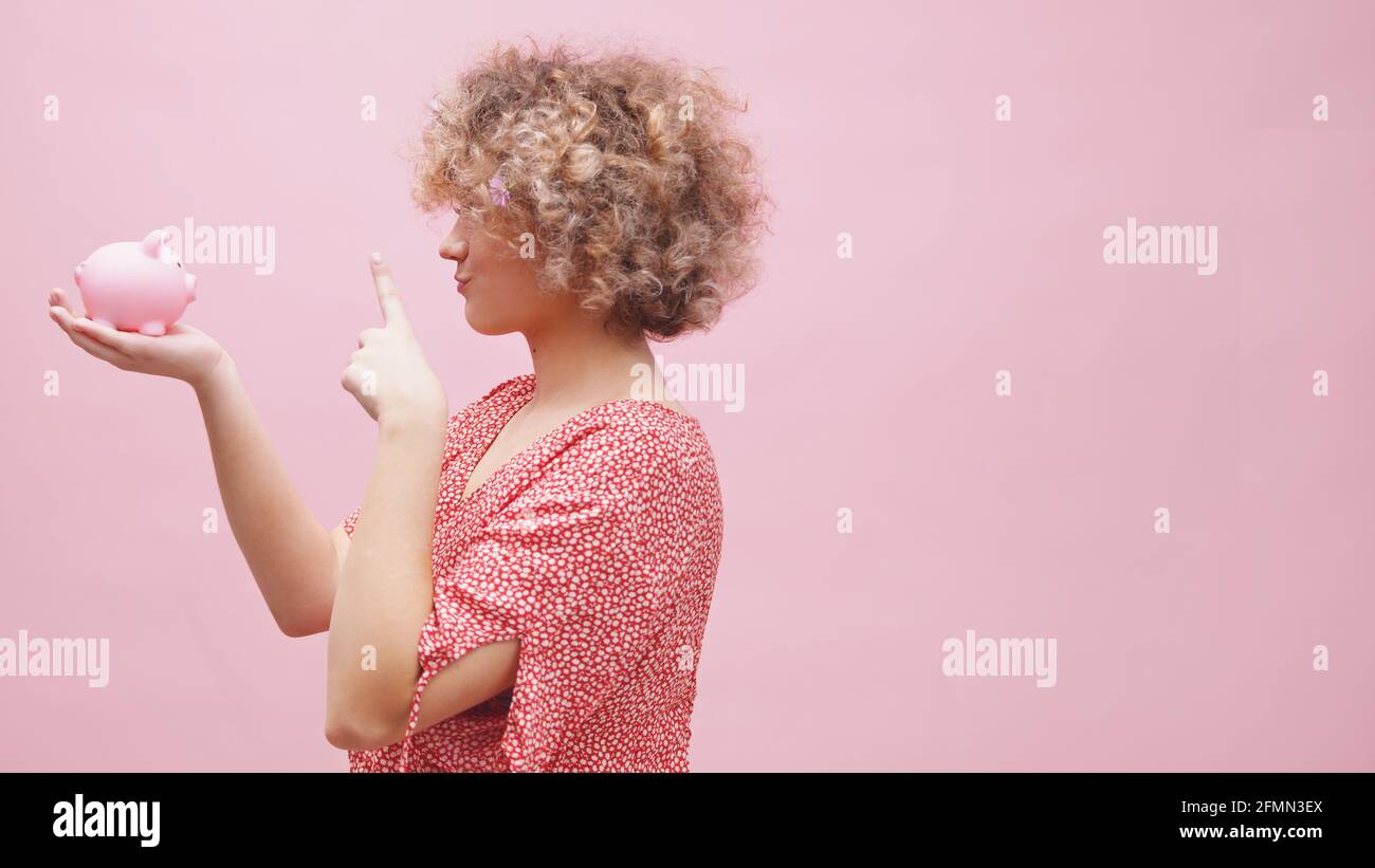 Jeune femme avec des fleurs dans les cheveux bouclés tenant la banque de porc et faisant de la magie avec un doigt pour l'épargne de croître. Photo de haute qualité Banque D'Images