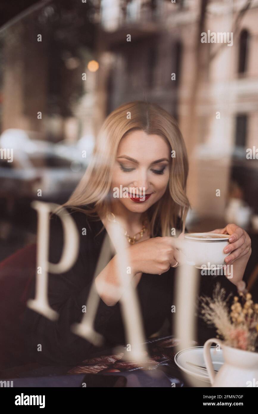 Bonne femme blonde dans un café avec une tasse de café aromatique, photo à travers la fenêtre de verre. Mise au point sélective douce, bruit artistique. Banque D'Images