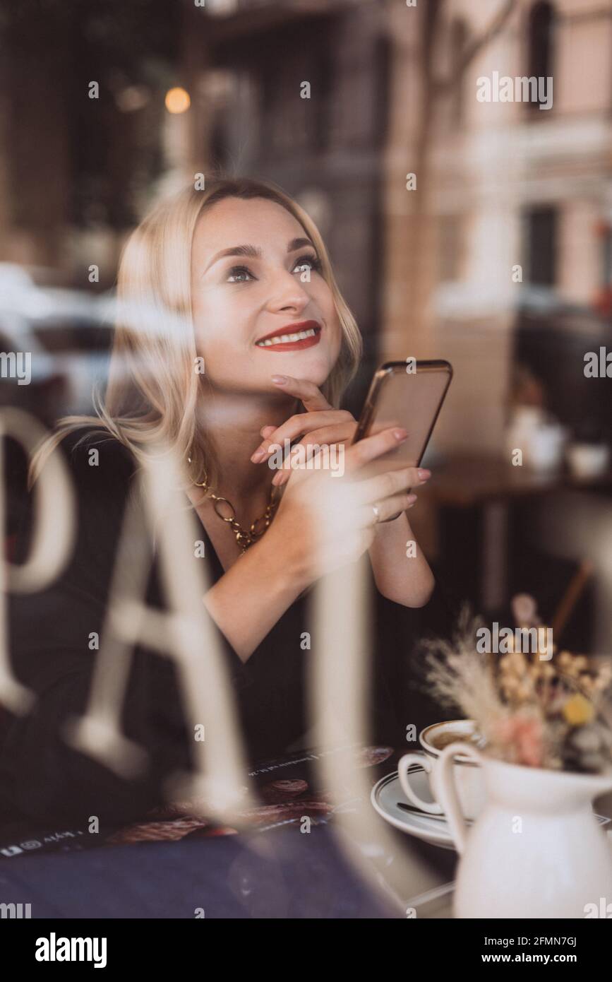 Bonne femme blonde dans un café avec une tasse de café aromatique et un smartphone dans ses mains, photo à travers la fenêtre de verre. Mise au point sélective, artiste Banque D'Images