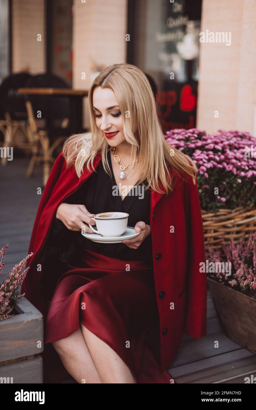 Belle jeune femme blonde dans une tenue d'automne tendance dans la couleur  bordeaux de marsala avec une tasse de café sur les marches d'un café. Mise  au point sélective douce. AR Photo