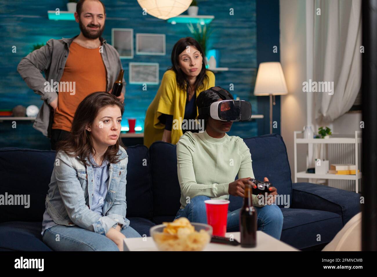 L'Afrique fait l'expérience de la réalité virtuelle jouant à des jeux vidéo pendant que ses amis sont en train de socialiser. Groupe de course mixte de personnes qui traînaient ensemble en s'amusant tard dans la nuit dans le salon. Banque D'Images