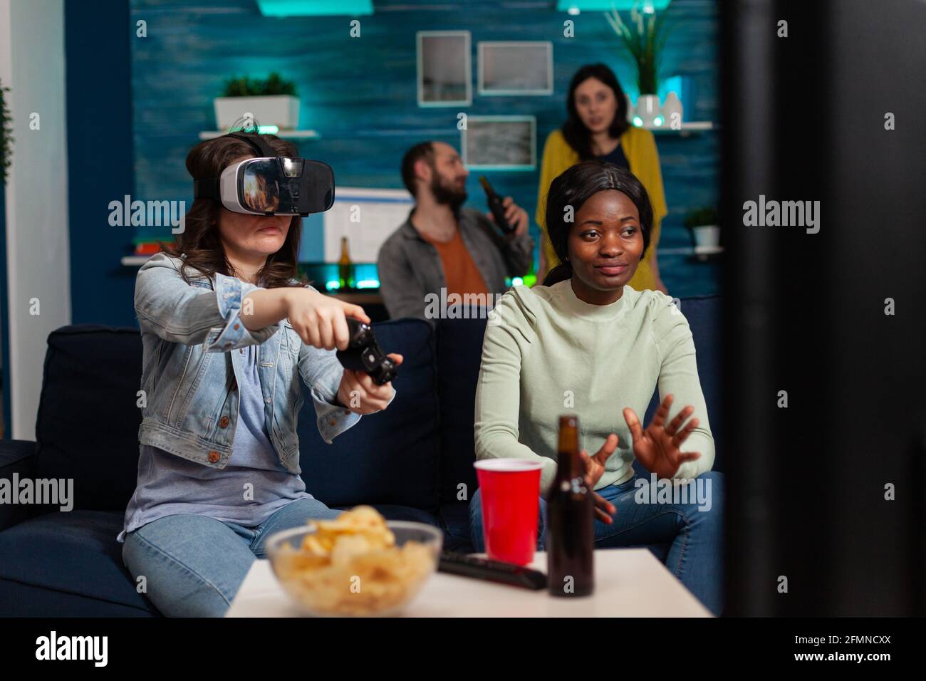 Une femme africaine qui applaudisse à ses amis lors d'un concours de jeux vidéo portant des lunettes de réalité virtuelle sur un canapé, s'amusant à l'aide d'une manette sans fil. Groupe ethnique mixte de personnes socialisant. Banque D'Images