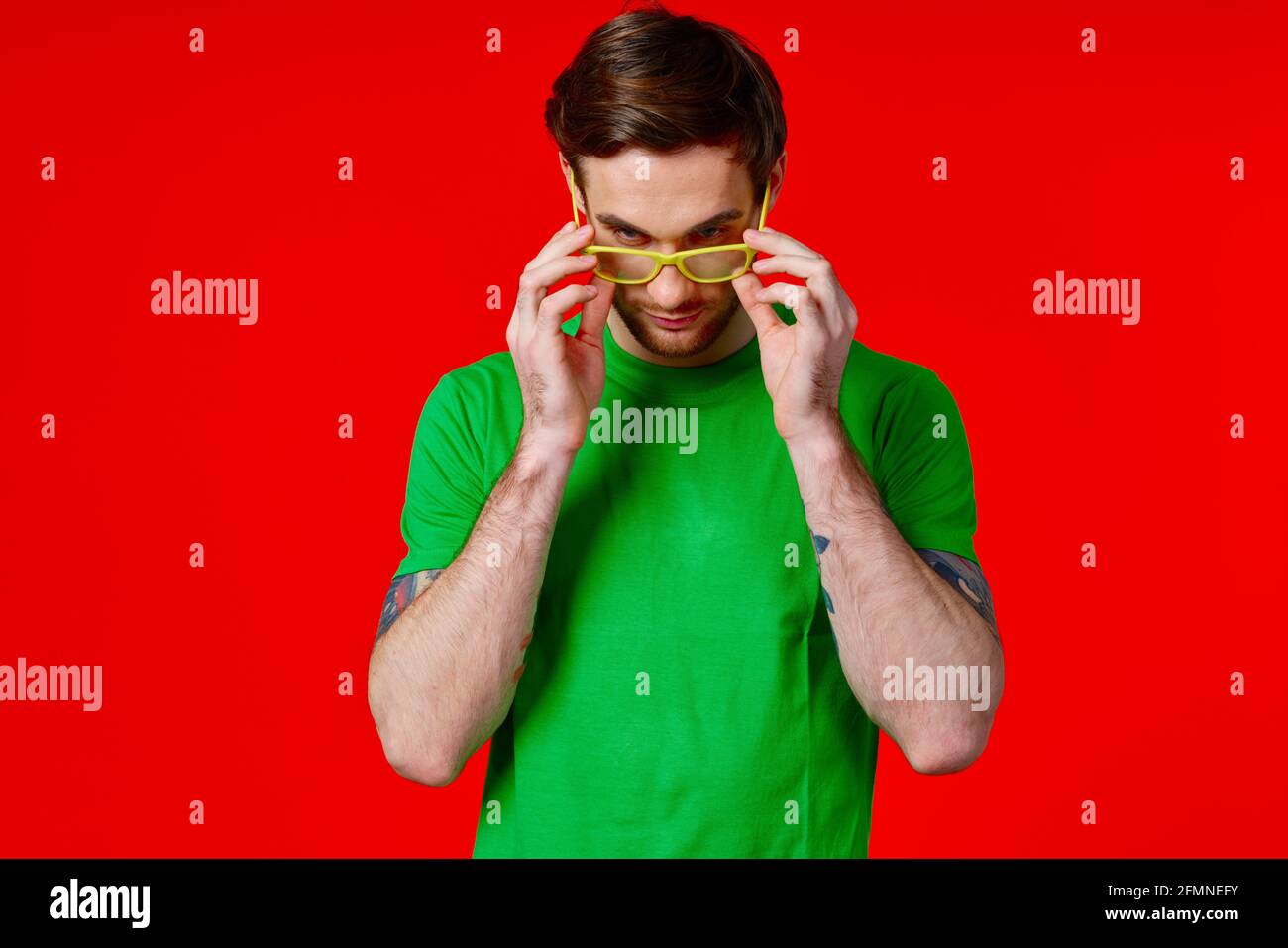 un homme dans un t-shirt vert avec des tatouages dans son mains portant des lunettes sur fond rouge Banque D'Images