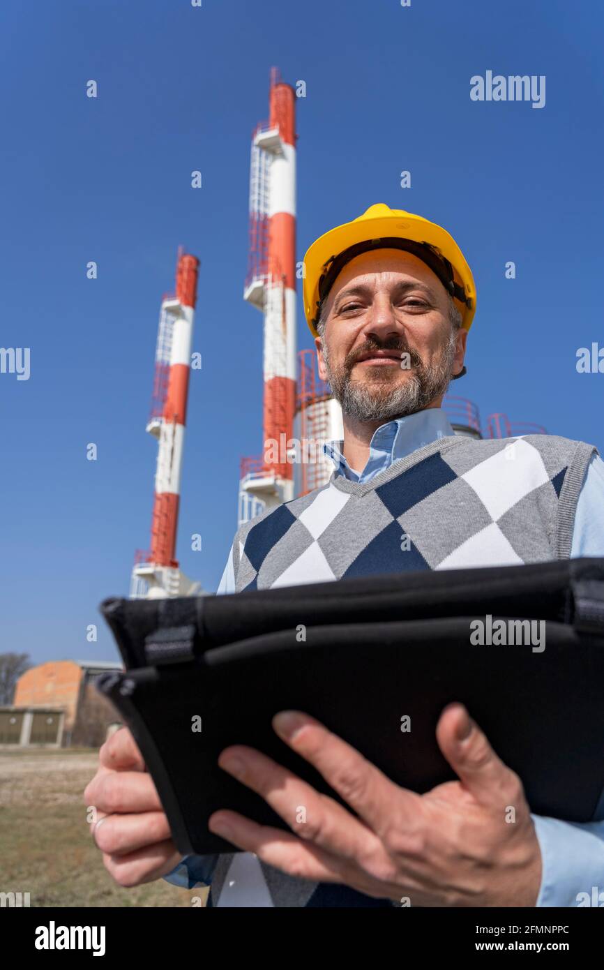 Homme d'affaires souriant dans Yellow HardHat tenant la tablette numérique et se tenant contre les cheminées de centrale électrique et le fond bleu ciel. Banque D'Images