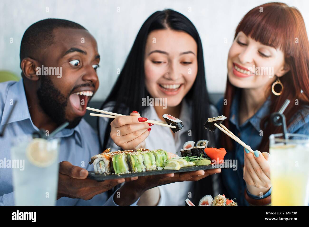 Trois jeunes gens multiethniques, enthousiastes et affamés, mangeant des petits pains à sushis japonais au restaurant avec bonheur et joie. Gros plan. Concept de réunion d'amis et de sushi Time Banque D'Images