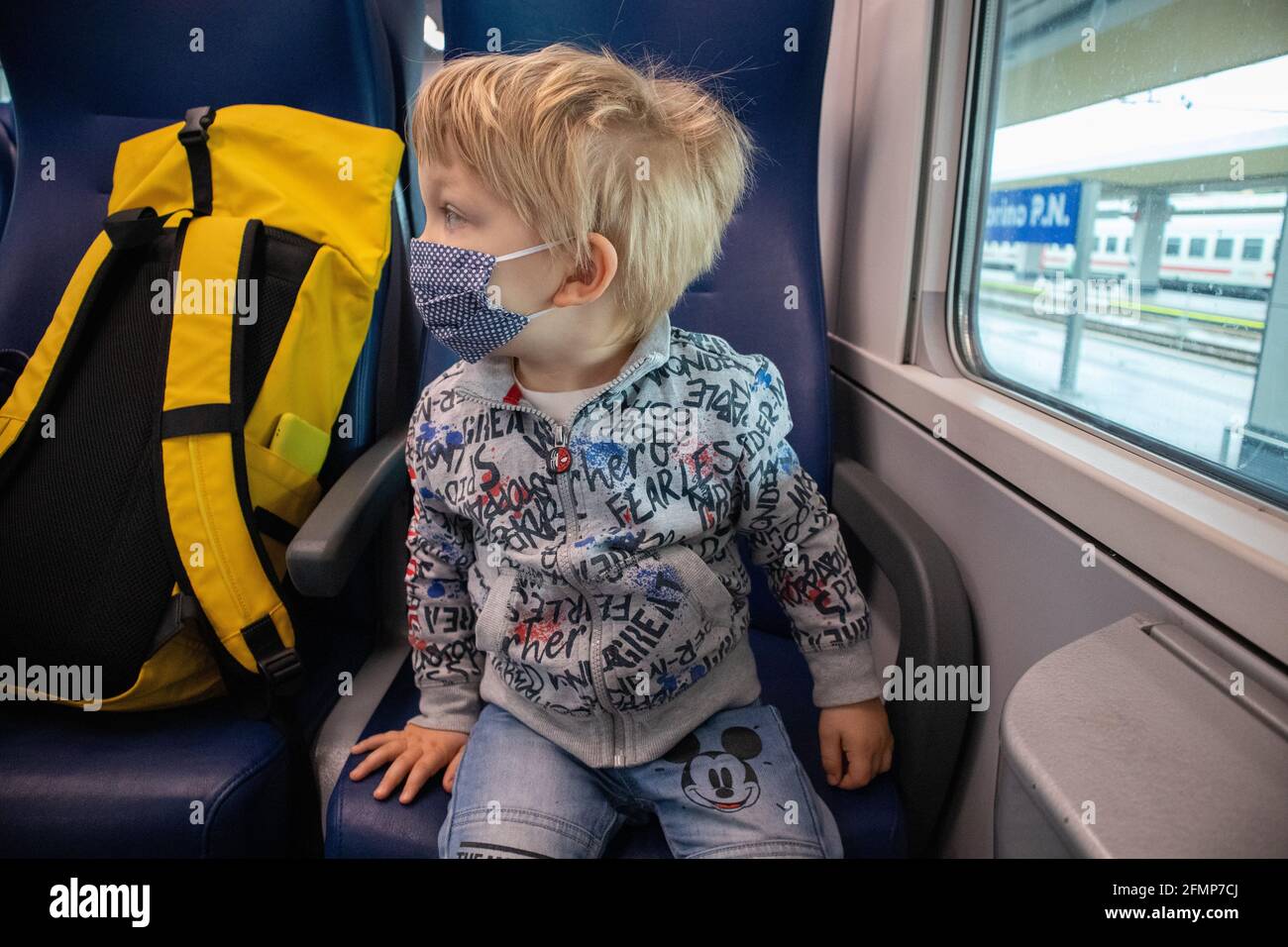 Un enfant blond avec des yeux bleus et un masque, près d'un sac à dos jaune, à bord d'un train régional en Italie Banque D'Images