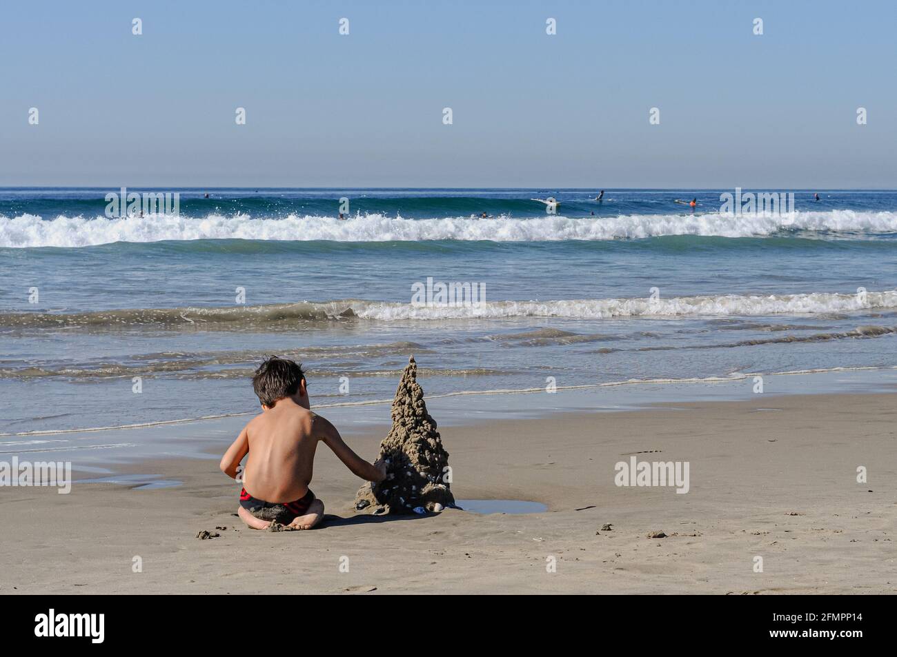 Un petit garçon créant un château de sable à lui seul à Venice Beach, Californie. Il a été pris dans l'après-midi. Les dudes surfeurs peuvent être vus en arrière-plan. Banque D'Images