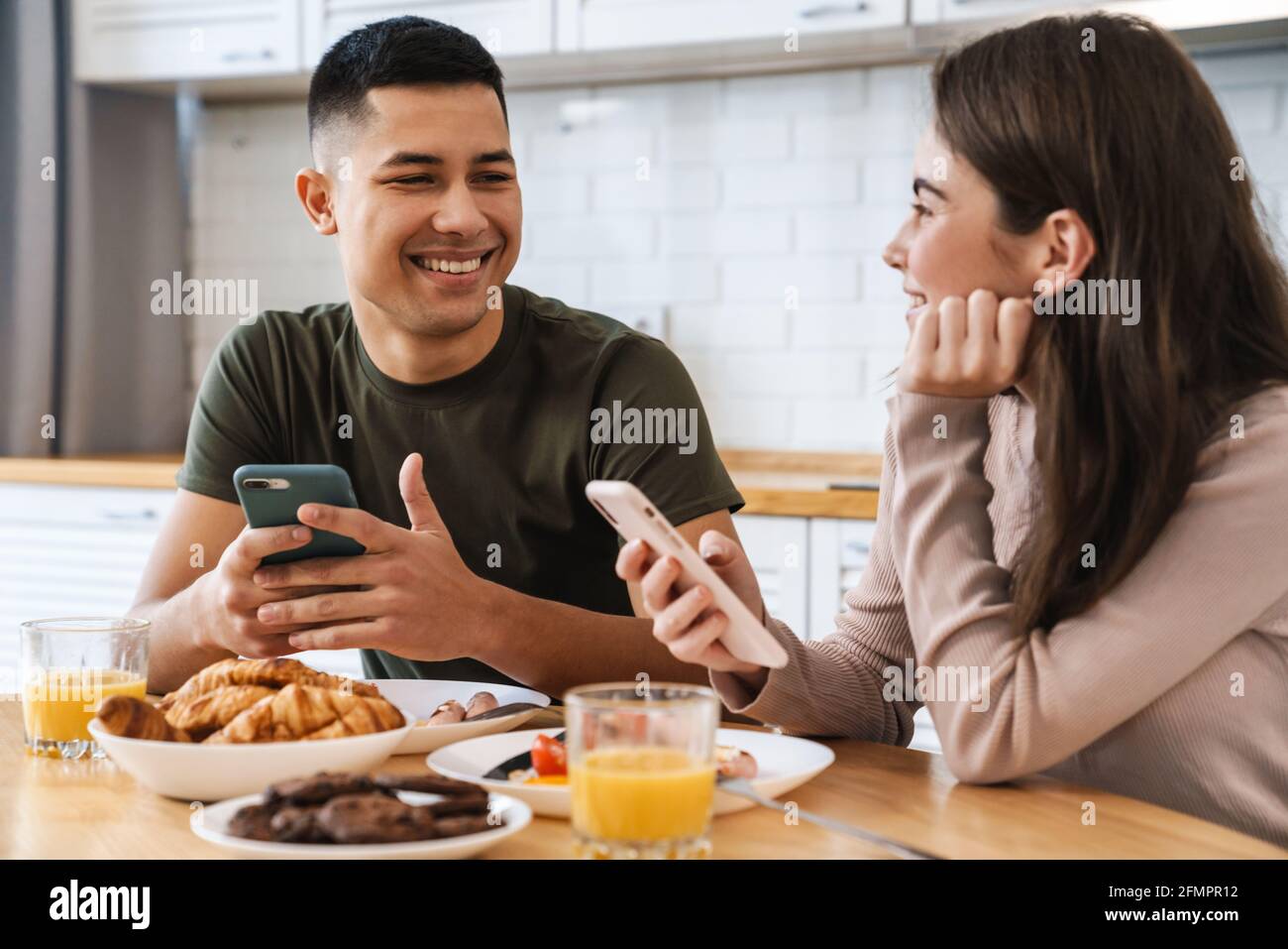 Portrait d'un couple souriant utilisant un smartphone et parlant tout en prenant le petit déjeuner dans une cuisine confortable à la maison Banque D'Images