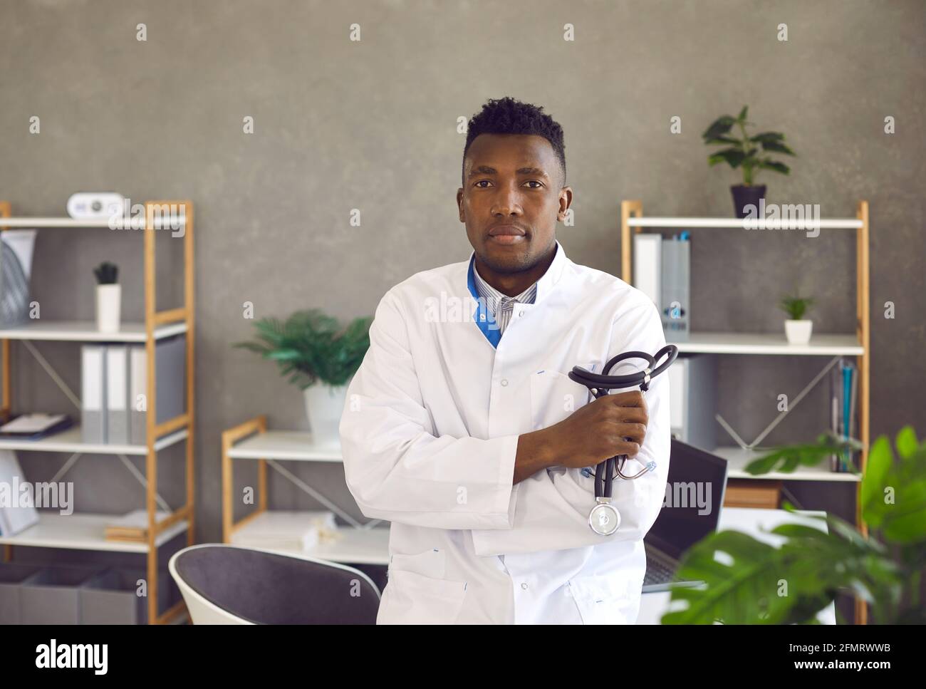 Jeune médecin noir debout dans le bureau médical, tenant le stéthoscope et regardant la caméra Banque D'Images