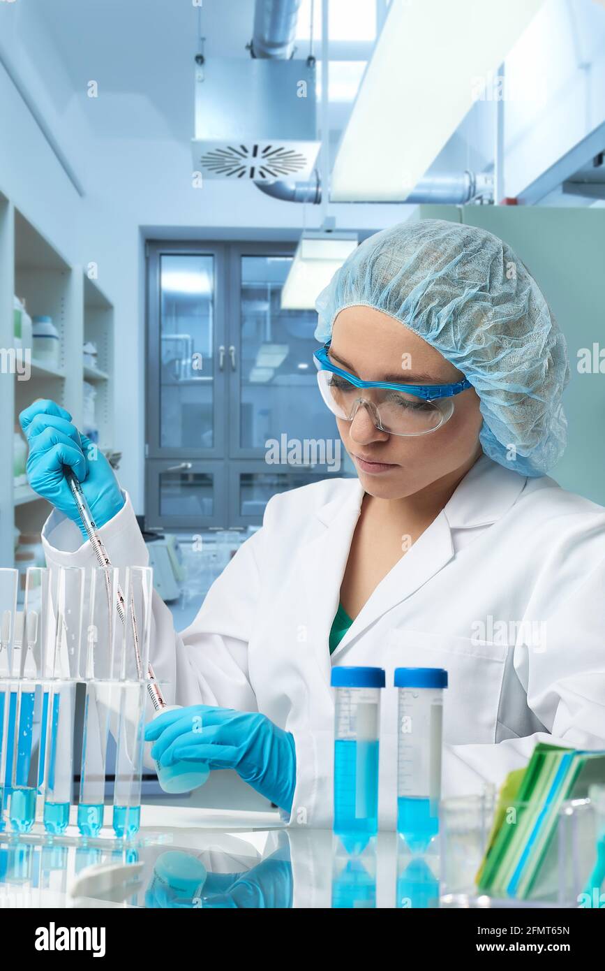 Pharma femme TECH travaille en laboratoire. Jeune femme caucasienne dans des gants de protection, un chapeau et une blouse blanche échantillon de pipettes dans un flacon. Intérieur du laboratoire Banque D'Images